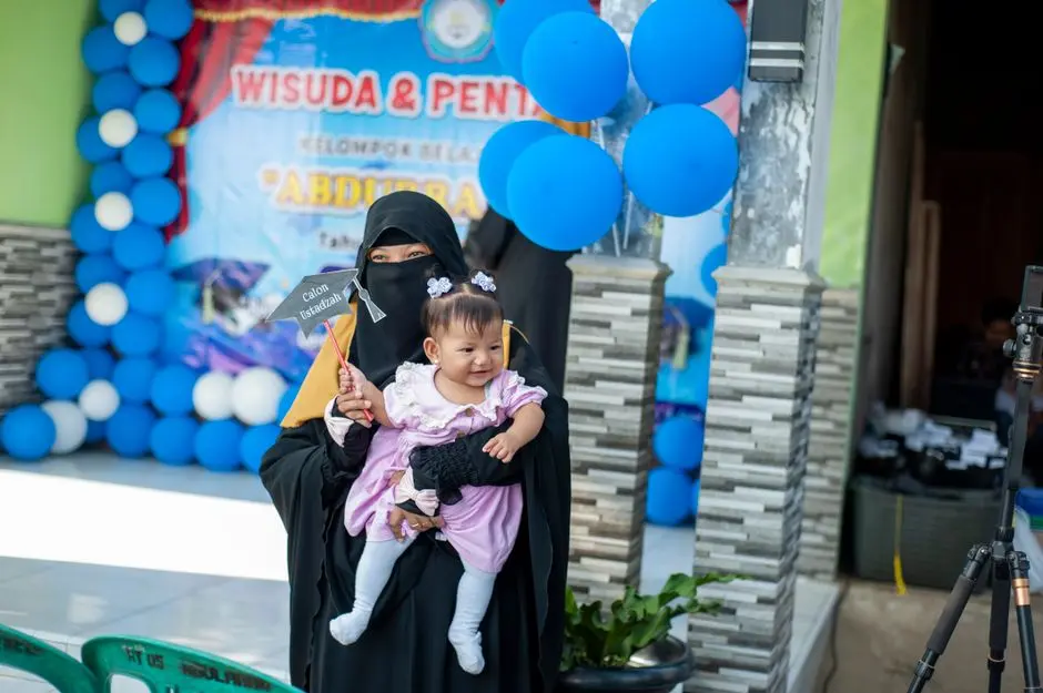 Ibu menggendong balita sambil tersenyum di teras rumah